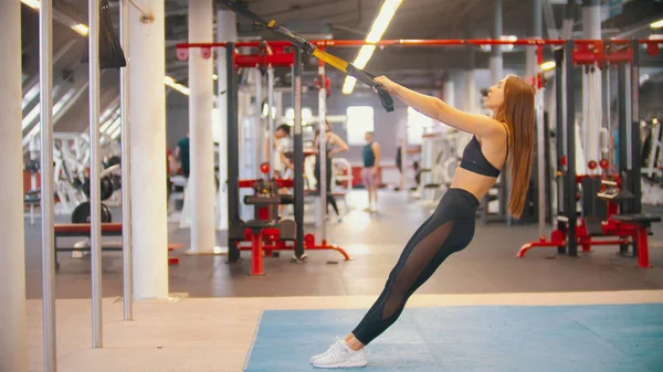 Una mujer deportiva en entrenamiento de ropa deportiva en el gimnasio, sujetando las asas y apoyándose hacia atrás —  Fotos de Stock