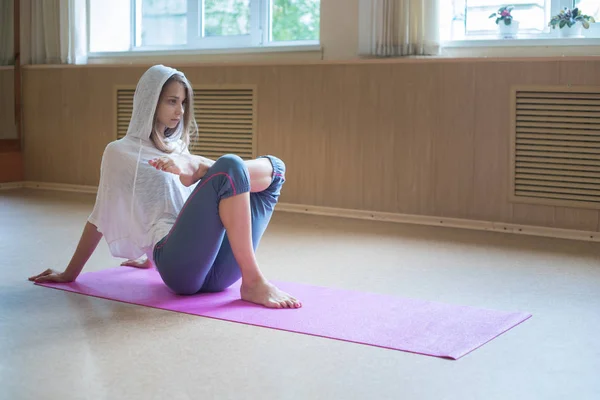 Fiatal karcsú nő, szőke haja ül a jóga mat és stretching lábát-elhelyezés egy lábát a térdét — Stock Fotó
