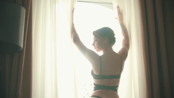 Jovem mulher em um sutiã preto sexy e calcinha em pé na frente da janela no quarto de hotel — Vídeo de Stock