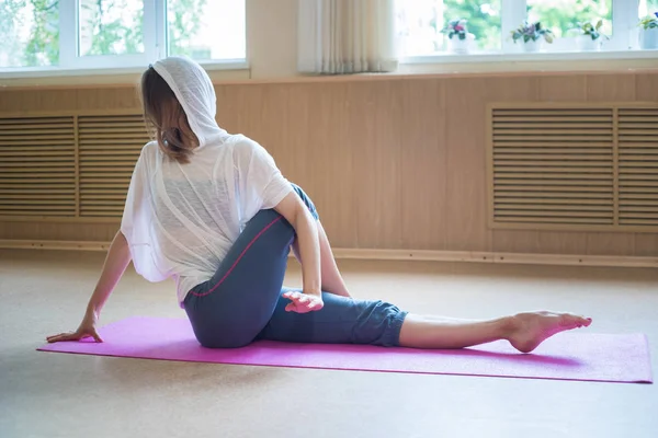Ung smal flexibel kvinna i huven sitter på yogamattan och stretching benet — Stockfoto