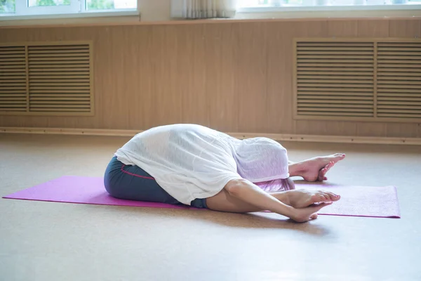 Jonge slanke vrouw zittend op de yoga mat en buigen naar voren naar haar tenen leunend op de benen — Stockfoto