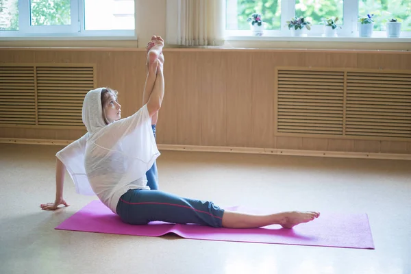 Junge Frau mit weißer Kapuze sitzt mit erhobenem Bein auf der Yogamatte - hält den Fuß und beugt ihn zur Seite — Stockfoto