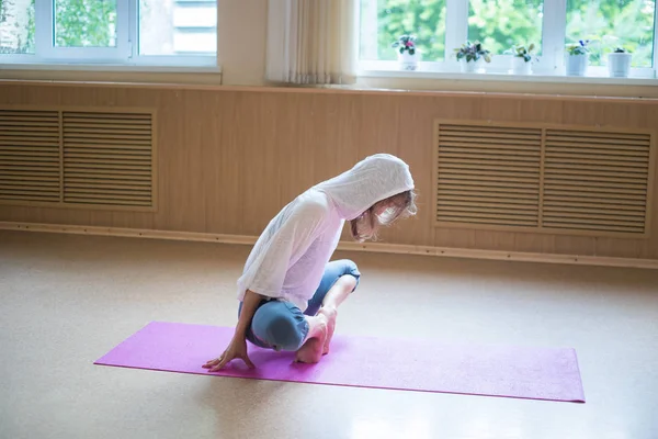 Jonge slanke vrouw in witte kap zittend op de yoga mat in lotuspositie leunend op haar tenen — Stockfoto