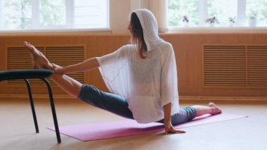 Genç güzel kadın yoga mat üzerinde oturan bir bölünmüş performans - bir sandalye kullanarak bacak germe egzersizleri yapıyor - dans stüdyosu