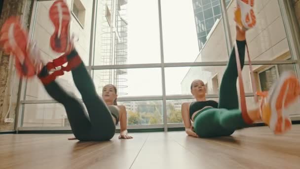 Dos mujeres sanas entrenando sus piernas usando una correa de estiramiento entre los tobillos — Vídeos de Stock