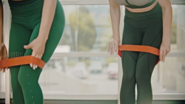 Dos mujeres con leggins verdes entrenando en el gimnasio, colocando una correa de estiramiento entre sus muslos — Vídeo de stock