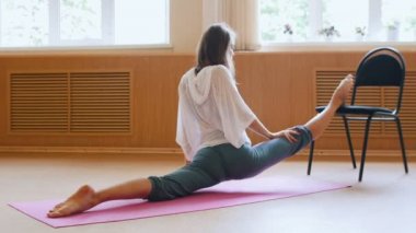 Bir bölünmüş performans yoga mat üzerinde oturan genç kadın - bir sandalye kullanarak bacak germe egzersizleri yapıyor