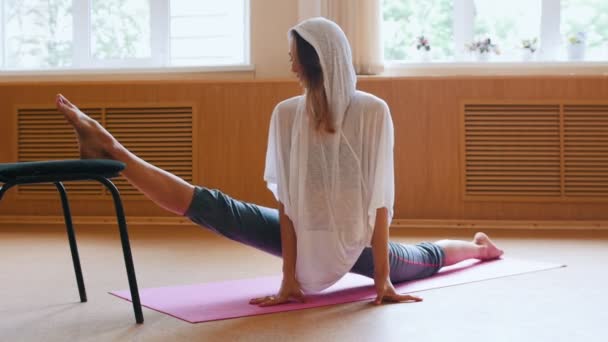 Giovane donna con cappuccio bianco seduta sul tappetino yoga che esegue una scissione - facendo esercizi di stretching delle gambe con una sedia — Video Stock