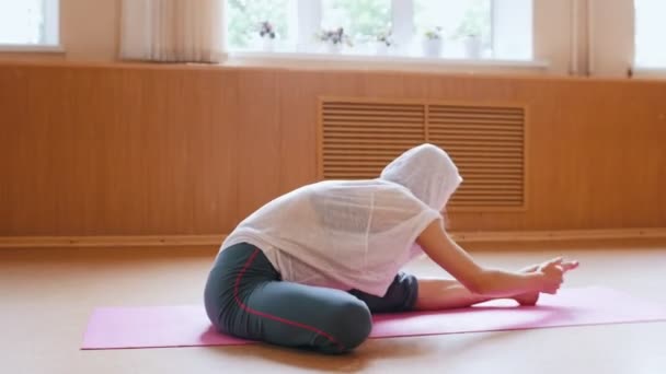 Mladá žena v bílém plášti, která sedí na rohoži jóga a nakláněla se dopředu na nohu — Stock video