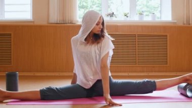 Bölünmüş yoga minderinde oturan genç ince kadın - bir ayak altında yardımcı stand kullanarak - öne eğilerek