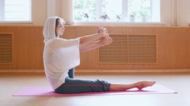 Yoga minderin üzerinde oturan kadar ısınma beyaz kapüşonlu genç ince kadın - ayak kapma ve bacak egzersizleri yapıyor