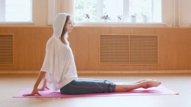 Beyaz kapüşonlu genç ince kadın yoga minderinin üzerinde oturuyor ve öne doğru onun tokalarını çekerek