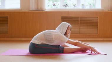 Yoga minderi üzerinde oturan ve öne eğilerek beyaz kapüşonlu genç ince kadın