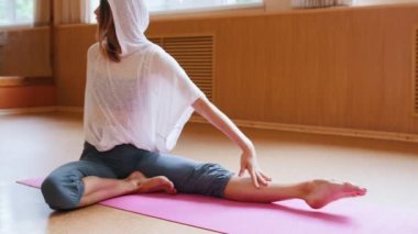 Yoga minderi üzerinde oturan ve bacak egzersizleri yapan beyaz kapüşonlu genç ince kadın - dans stüdyosu