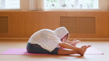 Yoga minderi üzerinde oturan ve öne eğilerek beyaz kapüşonlu genç ince kadın - ayaklara eğilerek