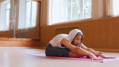 Yoga minderin üzerinde oturan ve öne eğilerek beyaz kapüşonlu genç ince kadın - eğilerek ve ayaklar üzerinde yere dokunarak