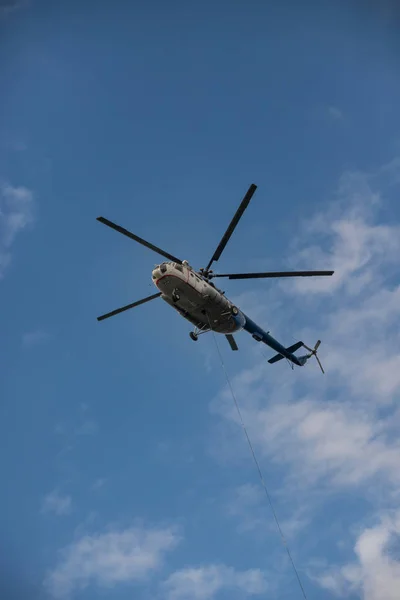 Mavi gökyüzünde uçan beyaz ve mavi bir helikopter - kablo helikopterdışında asılı — Stok fotoğraf