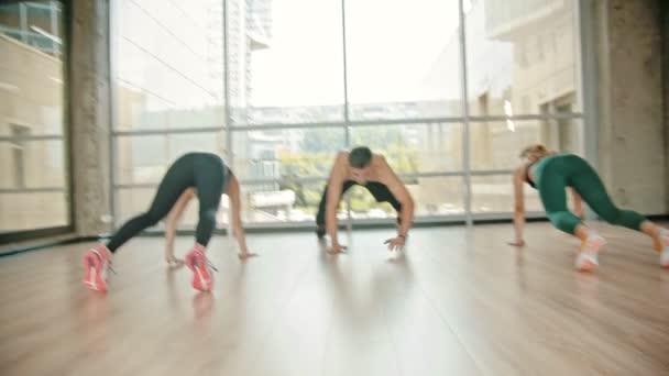 Фитнес-тренировка в студии - Две женщины и их тренер опираются на землю и ползают вперед - встают и прыгают — стоковое видео