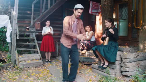 Muž, který tančí ruské lidové tance-ženy sedí na schodech u venkovského domu a pozoruje ho-žena hrající balalajka — Stock video