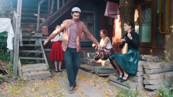 Un uomo barbuto che balla la danza popolare russa - donne sedute sulle scale vicino alla casa di campagna e che lo guardano - una donna che gioca a balalaika - ora legale — Video Stock