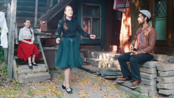 Una hermosa mujer bailando junto a la música cerca de la casa de campo - un hombre tocando balalaika — Vídeos de Stock