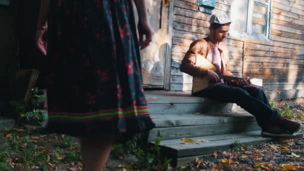 Žena tančící u hudby a baví se u domu-muž hrající balalajka-sluneční paprsky procházející stromy — Stock video