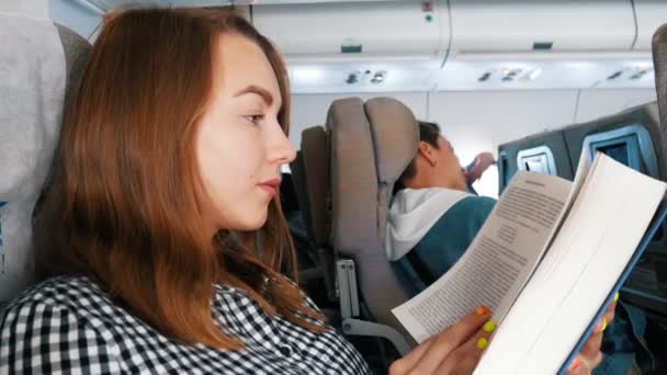 Una joven sentada en el avión leyendo un libro — Vídeos de Stock