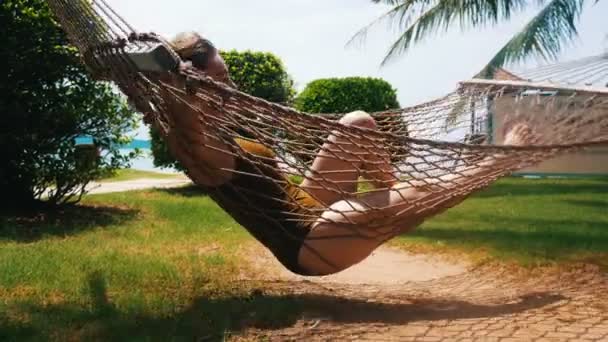 Een jonge mooie vrouw in zwempak liggend in de hangmat rond tropische planten en geniet van haar vakantie — Stockvideo
