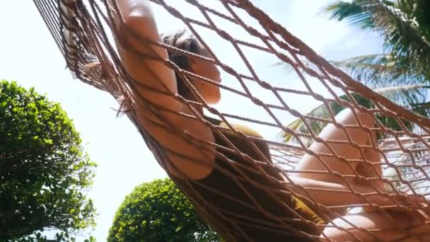 Uma jovem mulher bonita de fato de banho verde deitada na rede em torno de plantas tropicais e desfrutar de suas férias — Vídeo de Stock