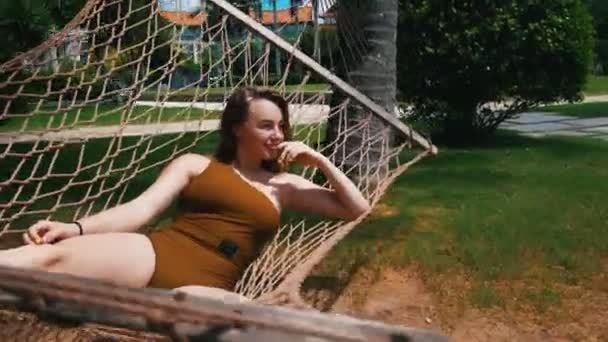 一个穿着单件泳衣的年轻漂亮女人躺在热带植物周围的吊床上休息 — 图库视频影像