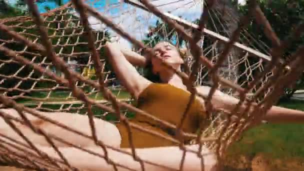 Молодая красивая женщина в цельном горчичном купальнике лежит в гамаке вокруг тропических растений и отдыхает — стоковое видео