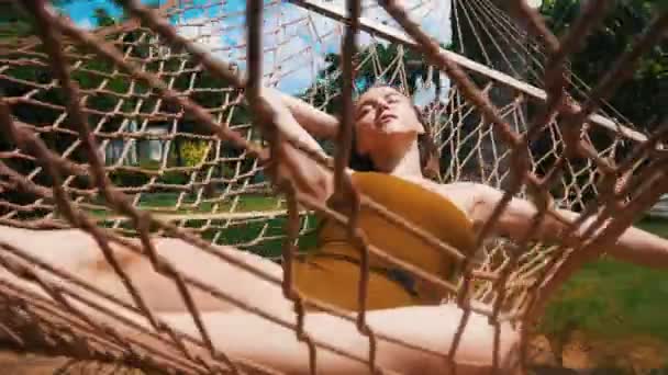 一个年轻的漂亮女人躺在吊床上，闭着眼睛围着热带植物休息 — 图库视频影像