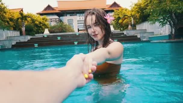 Молодая улыбающаяся женщина с мокрыми волосами ведет своего парня в бассейн за руку — стоковое видео