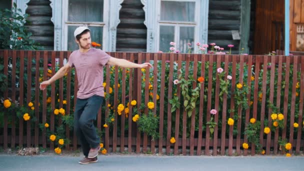 白い帽子をかぶった男が村の花でフェンスの前でロシアの民族舞踊を踊る — ストック動画