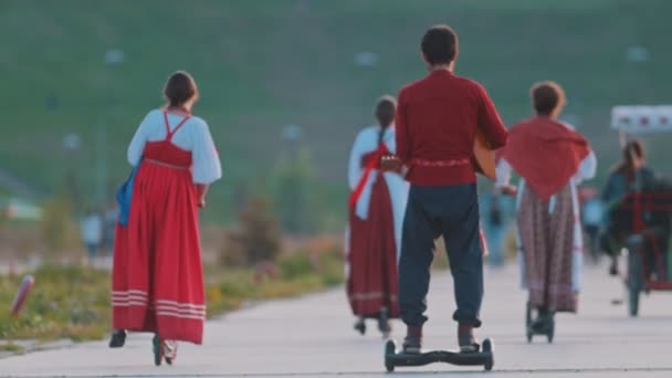 Personas en la música folclórica rusa caminando hacia el río en el hoverboard y un scooter — Vídeo de stock