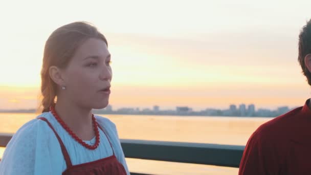 Lidé v ruských lidových kostýmech stojí na nábřeží během západu slunce a zpívají moderní budovy na pozadí — Stock video
