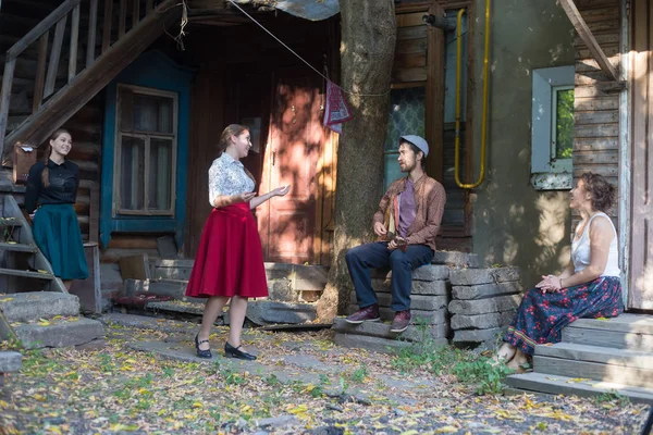 Company of people in russian clothes standing by the country house and talking - a man playing balalaika