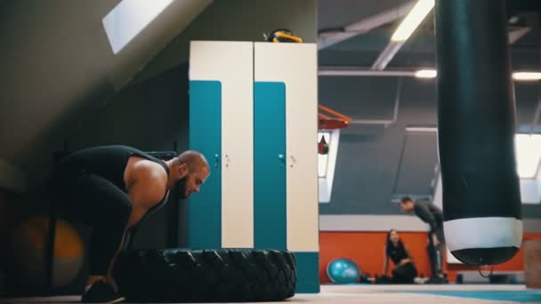 Een bebaarde man bodybuilder flipt een band op de vloer over en over in de sportschool-een andere mensen zittend op de vloer en praten — Stockvideo