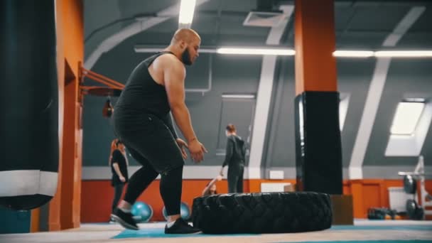 Ένας μουσάτος άνθρωπος bodybuilder γυρίζει ένα λάστιχο στο πάτωμα πάνω και πάνω στο γυμναστήριο-ένας άλλος άνθρωποι μιλούν στο παρασκήνιο — Αρχείο Βίντεο
