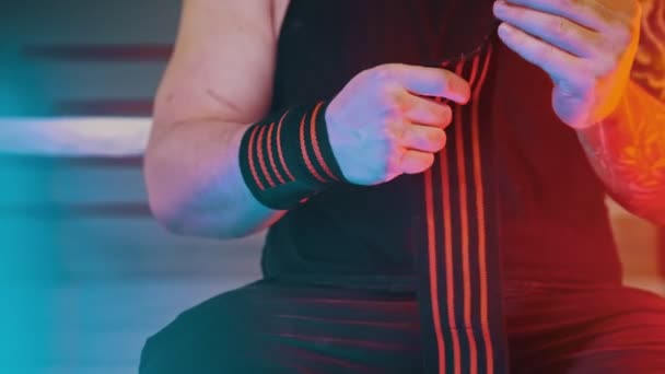 М'язистий чоловік з татуюваннями на руках наносить еластичну пов'язку на зап'ясті, сидячи в спортзалі в неоновому освітленні — стокове відео