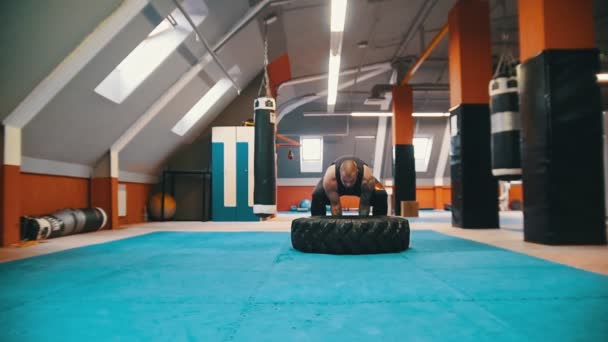 Ένα παχύ τατουάζ άντρας bodybuilder στροφή πάνω από το λάστιχο στο πάτωμα και μετακινώντας το με αυτή την κίνηση στο γυμναστήριο — Αρχείο Βίντεο