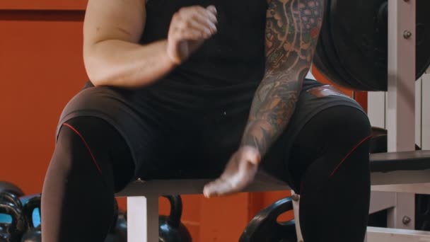 A muscular man with a tattoo sitting in the gym and clapping his hands covered with talcum — Stock Video