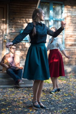 Kır evinin yanında dans eden uzun etekli iki kadın ve merdivenlerde balalaika oynayan bir adam.