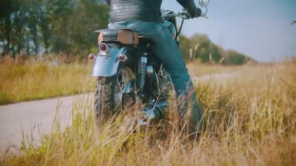 Ένας μοτοσικλετιστής αρχίζει να καβαλάει μια μοτοσικλέτα στο δρόμο που περιβάλλεται από το χωράφι με τη σίκαλη. — Αρχείο Βίντεο