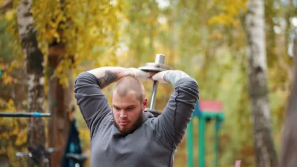 Bir adam vücut geliştirmeci arkasında dumbbell ile ellerini pompalama - açık spor zemin üzerinde eğitim — Stok video