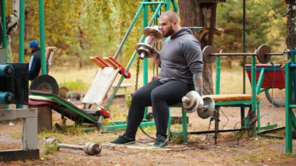 A man bodybuilder pulling the dumbbells in both hands - the dumbbells chained to the bench — Stock Video