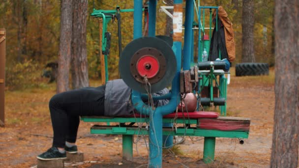 Bir adam vücut geliştirmeci bankta yatarken dambılile ellerini pompalama - sonbaharda açık spor zemin üzerinde eğitim — Stok video