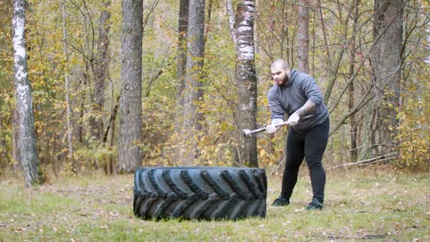 Um homem duro fisiculturista batendo o pneu do caminhão com um martelo de metal floresta de outono — Vídeo de Stock