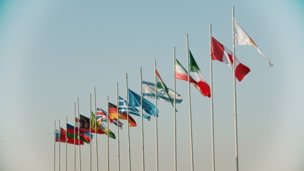 Banderas de los países del mundo que soplan en el viento en el fondo de la puesta del sol temprana en el cielo claro y hermoso — Vídeos de Stock