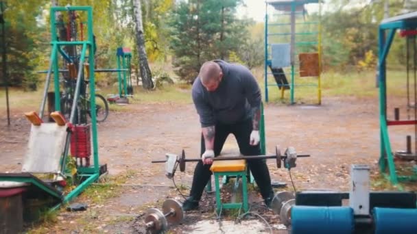 Een getatoeëerde man bodybuilder Pumping zijn handen met de dumbbells gemaakt van bakstenen en gewichten-training op het buiten sportterrein — Stockvideo